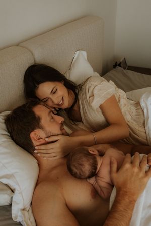 A tender family moment as parents cuddle with their newborn baby in bed, sharing laughter and joy together.