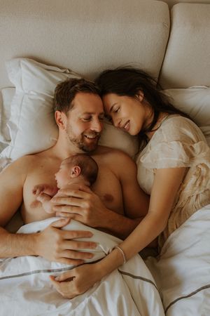 A tender family moment of a couple cuddling with their newborn baby in bed among white sheets and pillows.