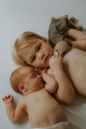 Toddler cuddling newborn baby sibling on white bed with stuffed animal toy.