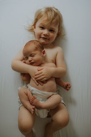 Toddler with blonde curly hair smiling while gently hugging a sleeping newborn baby on a white background.