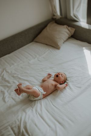 Baby wearing white diaper lies on white bedding while laughing joyfully on a bed with gray pillows.