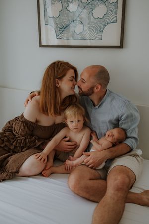 A couple shares a tender moment on a bed with their toddler and newborn baby, with wave art on the wall.
