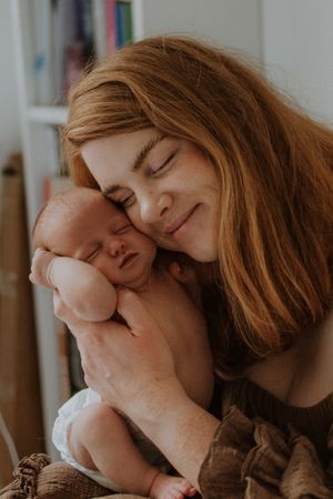 A red-haired mother smiles with eyes closed, gently pressing her cheek against her sleeping newborn baby.