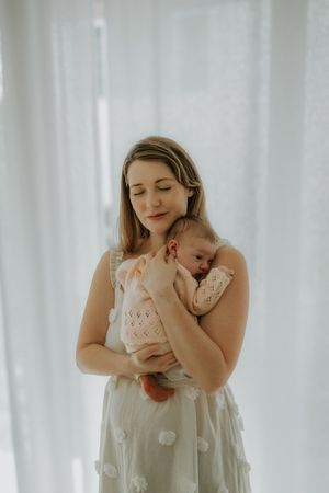 A tender moment between mother and baby in white lace dress against sheer curtains in a bright, airy setting.
