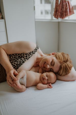 A smiling mother in leopard print top cuddles her sleeping newborn baby on a white bed.