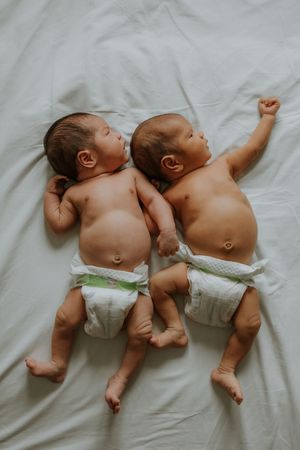 Two newborn twins lying side by side on a white bed, wearing diapers, in a peaceful resting pose.