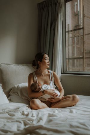 A tender moment captured in soft natural light as a mother nurses her baby while reclining on white bedding near a window.