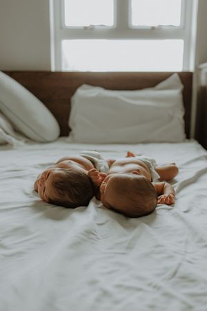 Two newborn twins lying side by side on a white bed, sleeping peacefully near a sunlit window.