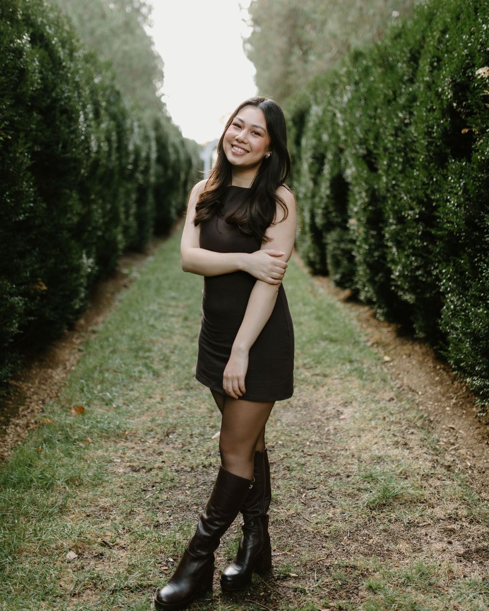 A person in a black dress poses gracefully on a tree-lined path surrounded by lush greenery.
