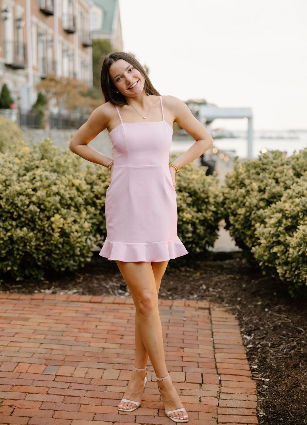 A person in a pink ruffled summer top poses playfully on a brick pathway next to landscaped shrubs.