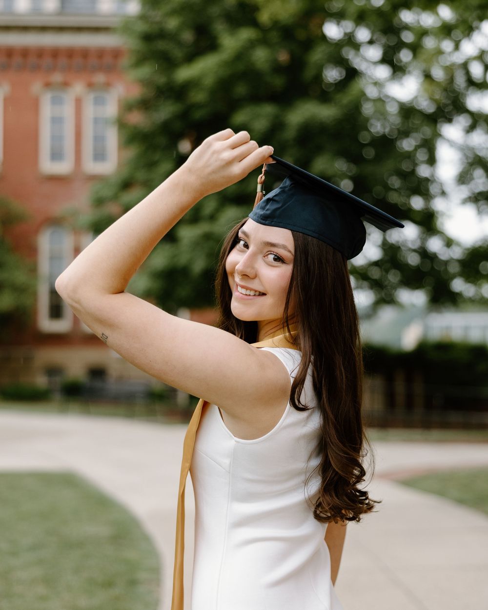 Graduate in white dress and black cap poses outdoors on campus with joyful expressions.