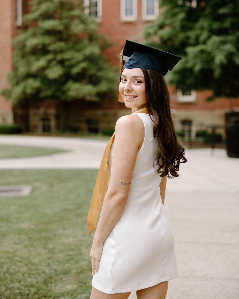 Graduate in white top and graduation cap poses outdoors on campus with brick buildings in background.