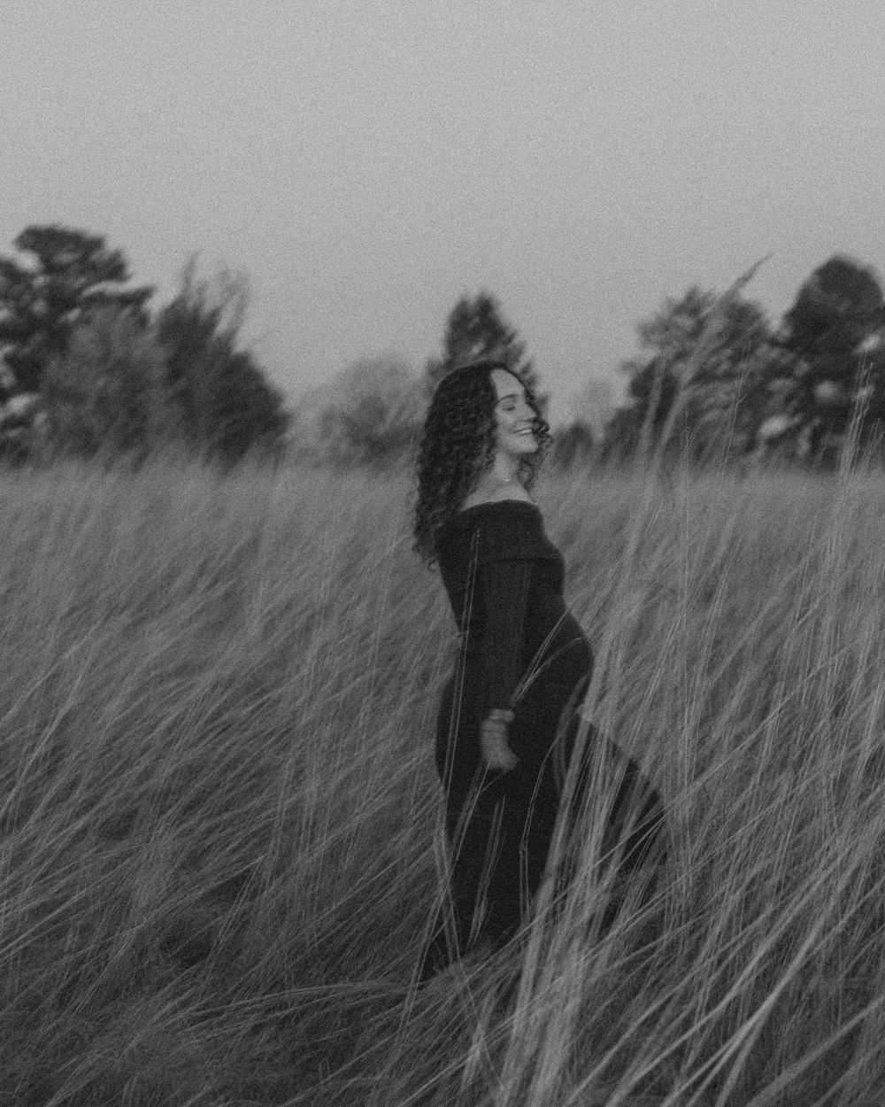 A silhouetted figure stands in tall grass against an overcast sky in a moody black and white landscape photograph.