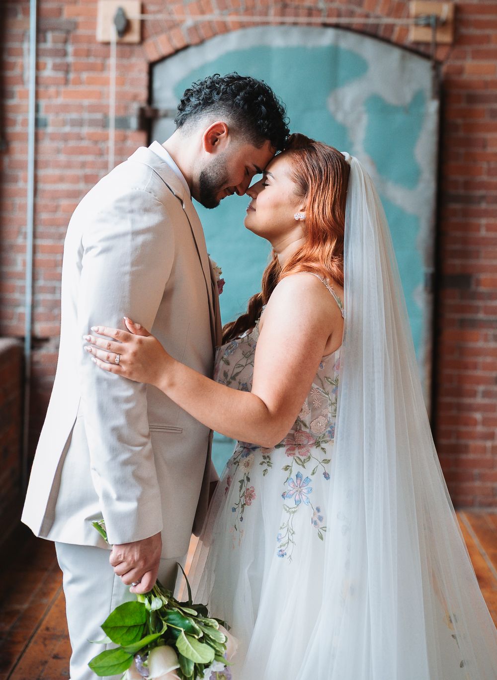 A bride and groom share a romantic forehead touch in a rustic brick venue, she in a floral gown with veil, he in a tan suit.