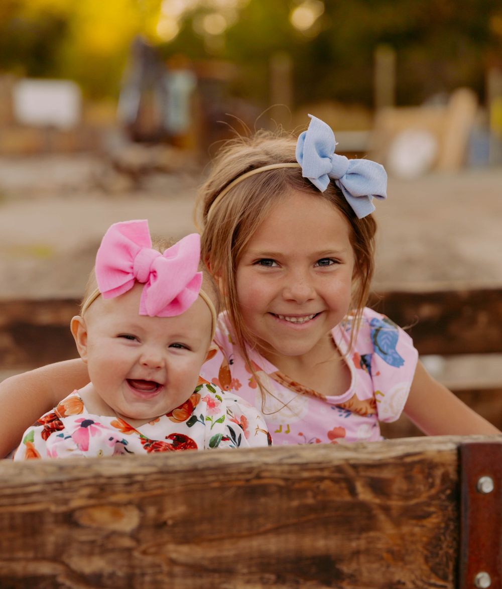 Children wearing floral outfits and bows peek over a wooden fence in an outdoor setting.