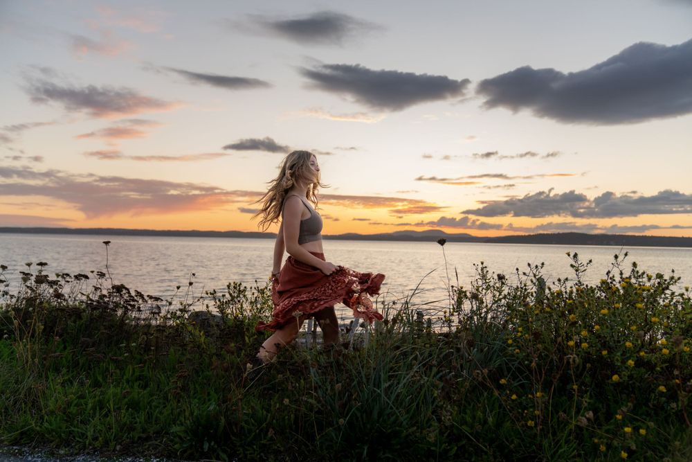 Silhouette of person in flowing dress dancing by ocean at sunset among wildflowers by Filling the Frame Photography's at Lopez Island in San