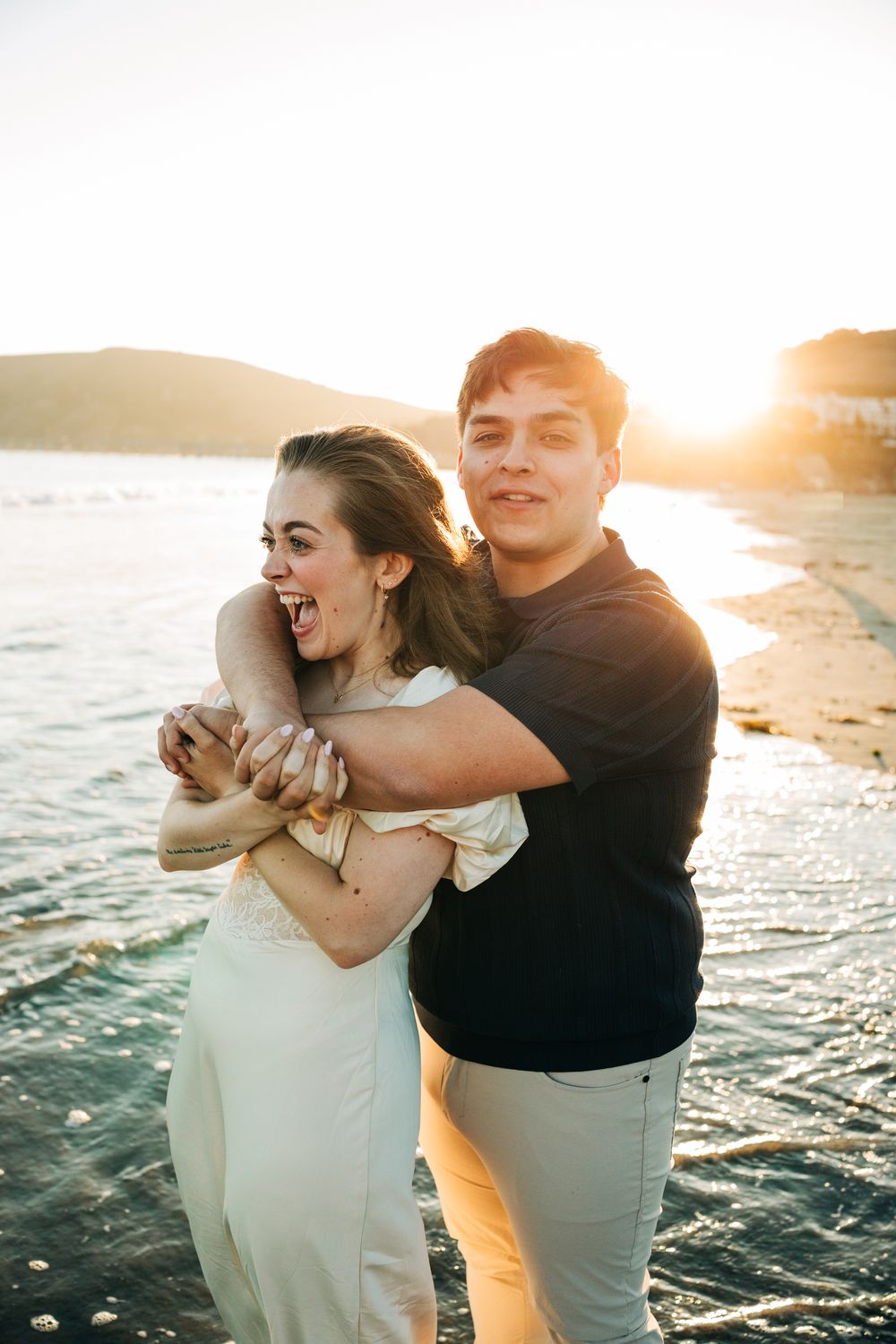 Engagement session in Avila Beach, CA. Man hugging his fiance from behind, in front of the sunset.