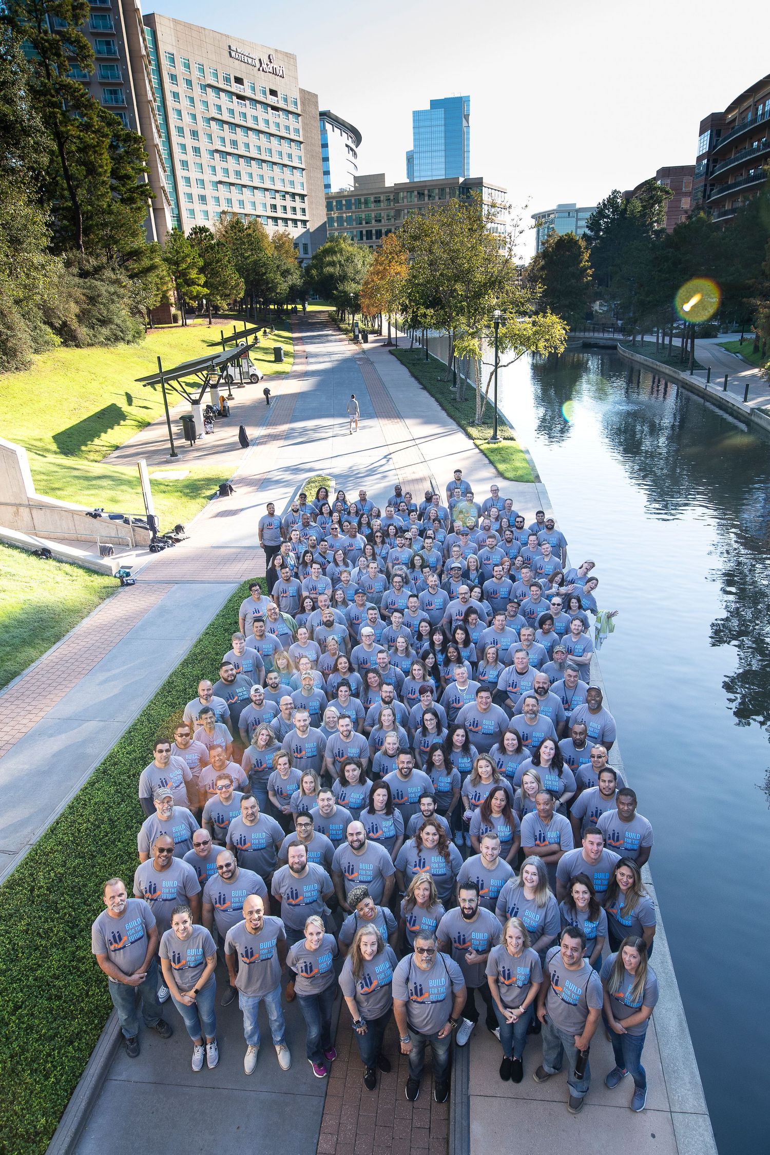 Large group of employees in matching gray shirts pose along a scenic urban waterway with city buildings in the background.