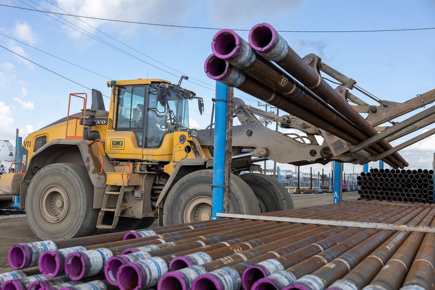 Yellow front loader transporting purple and black industrial pipes at an outdoor construction or oil field equipment yard.