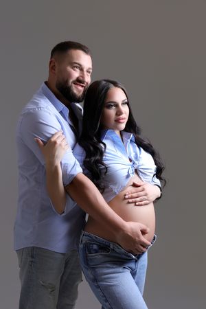 Couple embracing in casual attire during pregnancy photoshoot against neutral backdrop.