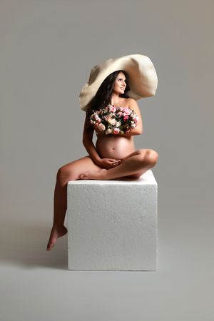 Artistic maternity pose on white block with floral crown and natural lighting.
