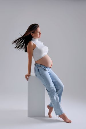 Pregnant woman in white crop top and blue jeans leaning on white cube with flowing dark hair on gray background.