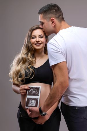 Couple holds ultrasound photos during pregnancy announcement photoshoot against gray backdrop.