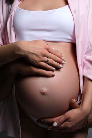 Close-up of hands gently cradling pregnant belly in white crop top and pink cardigan.