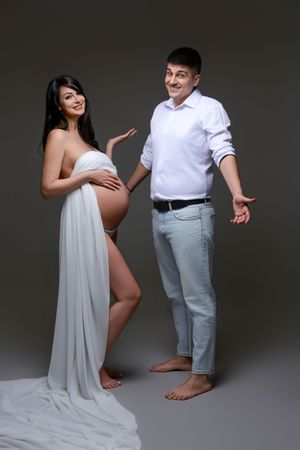Pregnant woman draped in white fabric poses with smiling man in white shirt and jeans against gray background.