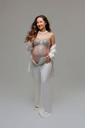 Pregnant woman in rhinestone bodysuit, white pants and sheer wrap poses gracefully against a gray background.