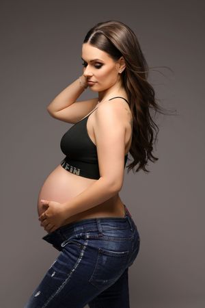 Maternity photo session showing pregnant woman in black top and jeans against grey background.