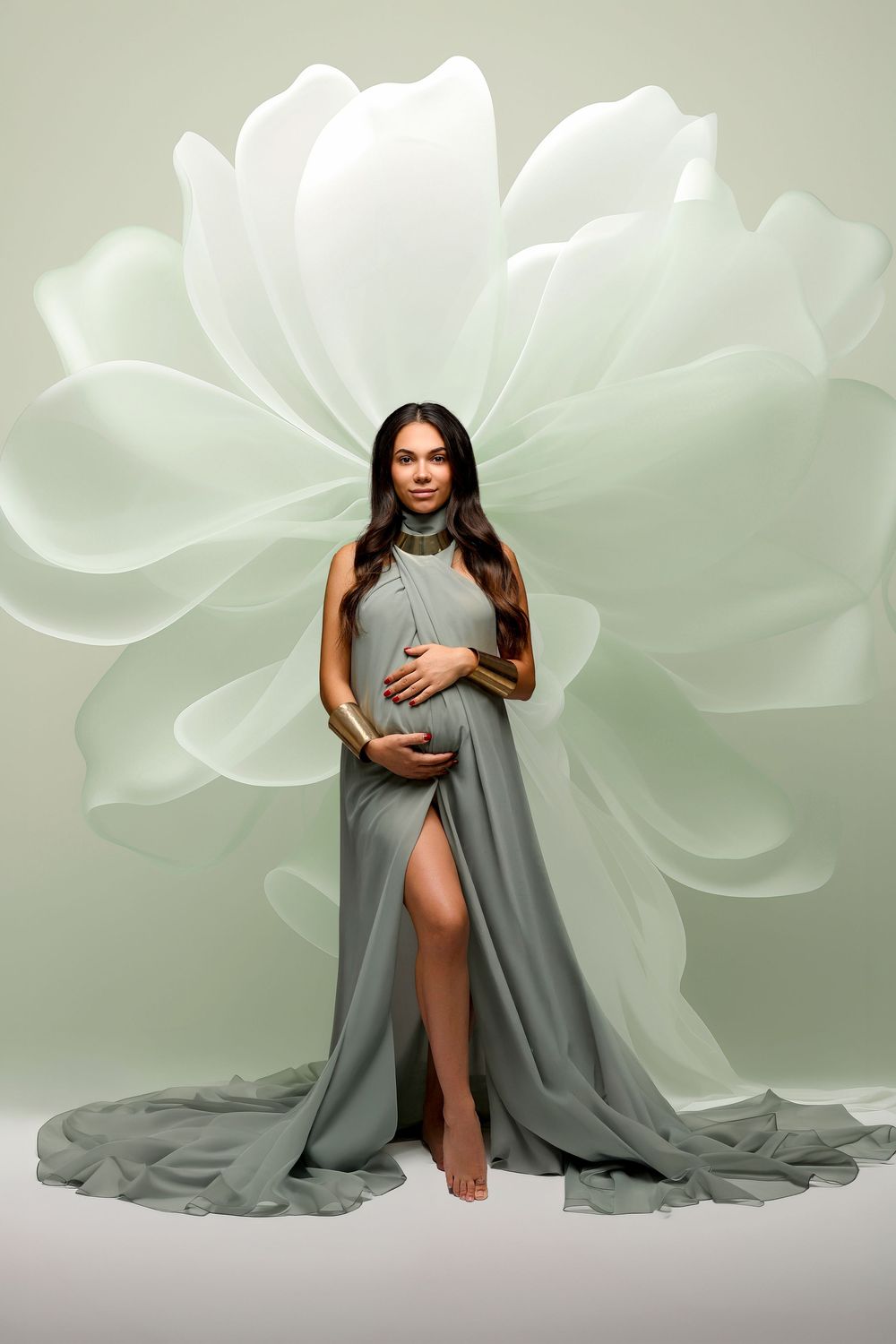 Pregnant woman in elegant sage green halter gown with flowing train, posing before large floral backdrop.