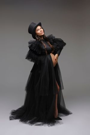 Pregnant woman in black tulle gown and top hat poses elegantly against gray background.