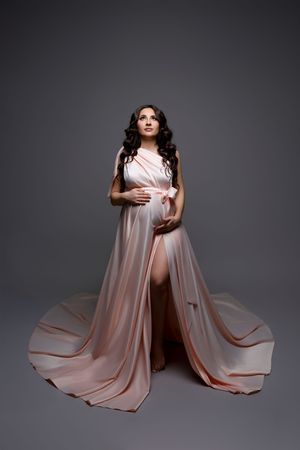 Pregnant woman in flowing blush pink gown with train poses gracefully in studio maternity portrait.