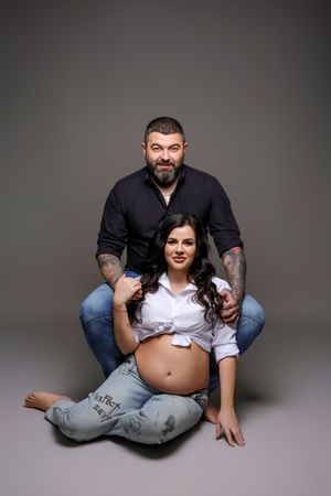 Pregnant woman in white shirt sits on floor with tattooed bearded man behind her, both smiling in studio photo.