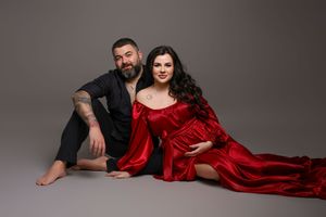 Pregnant woman in red silk gown poses with tattooed man in black outfit for maternity photo shoot.