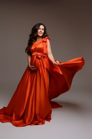Pregnant woman in flowing red satin gown posing gracefully in studio, fabric billowing elegantly around her.