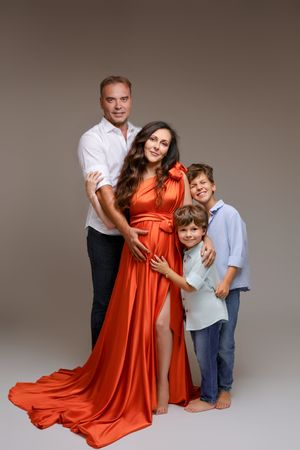 Pregnant woman in orange gown poses with husband and two sons in a warm studio family portrait.