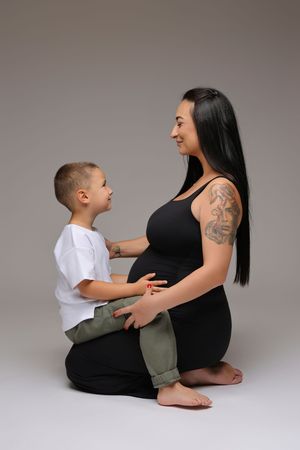 Pregnant woman with tattoos sits on floor smiling at young boy who touches her baby bump in studio.