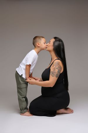 Pregnant woman in black outfit with tattoo kneeling while young boy in white shirt kisses her on grey background.