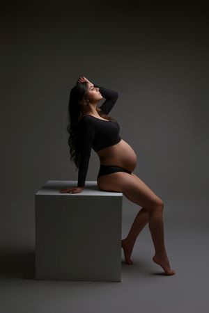 Pregnant woman in black crop top and underwear posing gracefully on white cube in a studio setting.