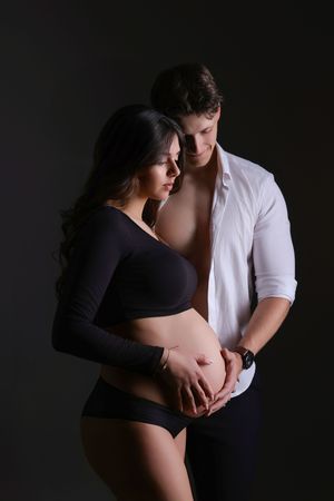 A couple tenderly cradles a pregnant belly together in a dark studio, the woman in black lingerie and man in an open white shirt.
