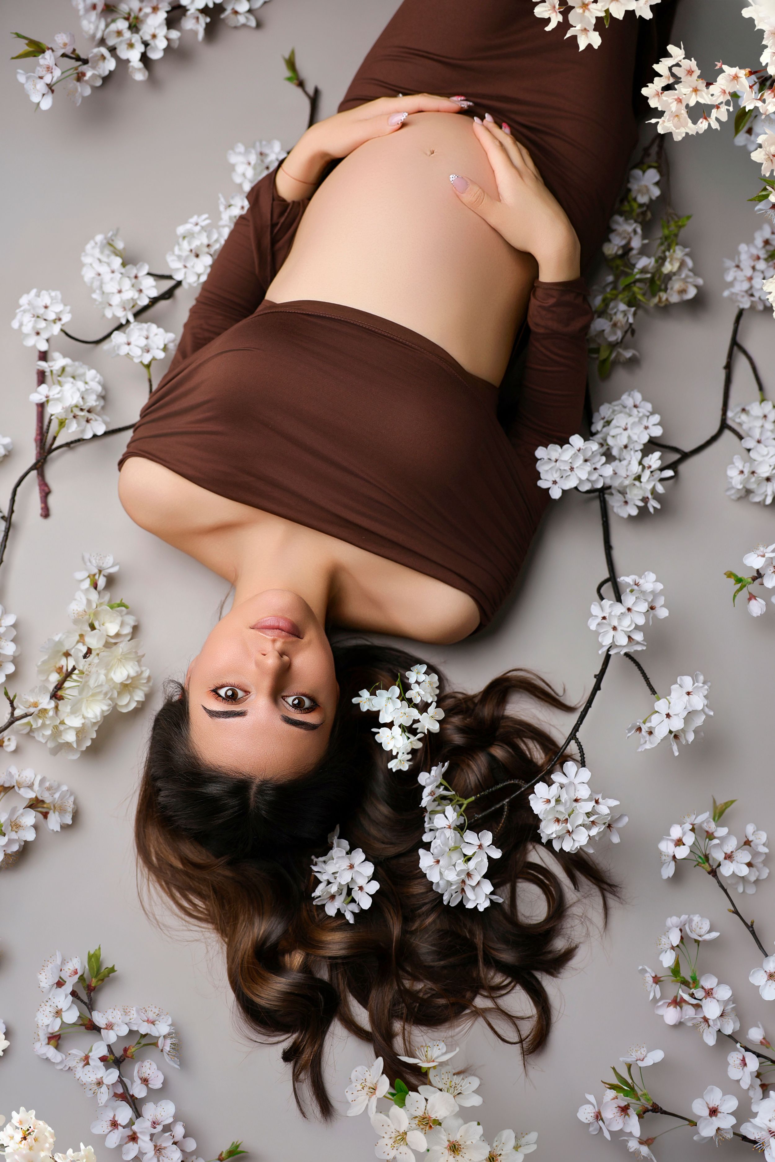 Pregnant woman in brown dress lying among white cherry blossoms, cradling her baby bump in a serene studio setting.