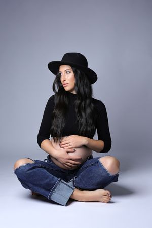 Stylish woman in black hat and turtleneck sitting cross-legged for maternity photo.