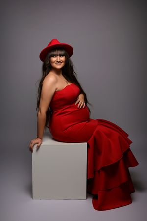 Elegant maternity studio session with woman in red dress posing on white cube.