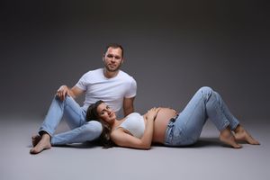 Pregnant woman lying on floor with partner in white tee and jeans posing together in studio photo shoot.