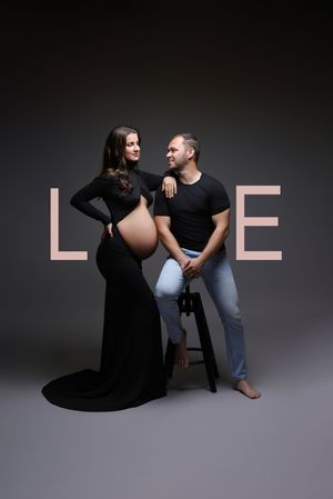 Pregnant woman in black gown poses with partner beside stool against dark backdrop with LOVE text.