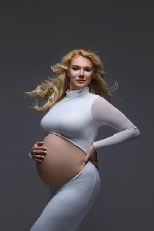Pregnant woman in white bodysuit cradling her baby bump, with flowing blonde hair against a gray background.