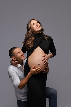 A loving couple poses for maternity photos with the partner embracing the pregnant belly in a black dress against gray backdrop.