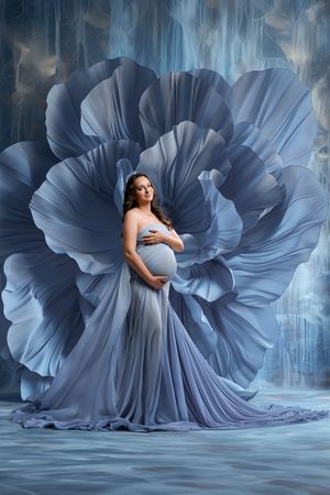 Pregnant woman in flowing blue gown poses gracefully against a large blue floral backdrop in an artistic maternity portrait.